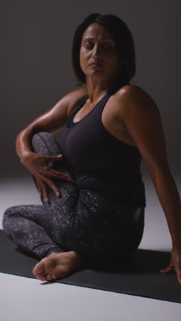 Vertical-Video-Studio-Shot-Of-Mature-Woman-Wearing-Gym-Fitness-Clothing-Sitting-On-Floor-Doing-Pilates-Stretching-Exercises